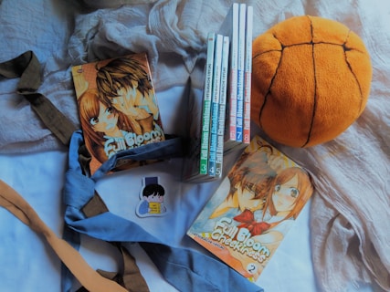 A collection of manga volumes titled 'Full Bloom Cheekiness' is arranged alongside a plush basketball. A few ties in shades of blue and brown are artfully placed. There is a sticker depicting a person reading a book nearby.
