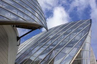 Modern architectural structures with large, curved glass panels against a blue sky with white clouds. The buildings appear to have a sleek, futuristic design with reflective surfaces and metallic support elements.