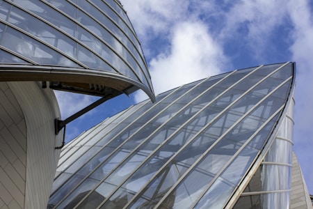 Modern architectural structures with large, curved glass panels against a blue sky with white clouds. The buildings appear to have a sleek, futuristic design with reflective surfaces and metallic support elements.