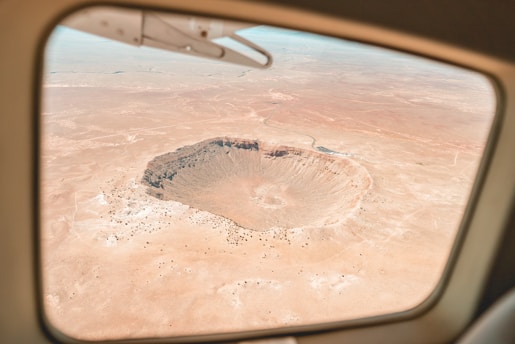 A large, circular meteor crater is visible through an aircraft window, situated in an expansive, arid desert landscape. The terrain is mostly barren with sparse vegetation, highlighting the crater's prominence.