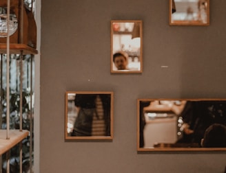 Multiple wooden-framed mirrors are mounted on a gray wall, reflecting a blurred section of a room and a person partially visible. The room appears dimly lit with warm, ambient lighting.