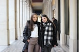 Two women stand side by side in a well-lit, elegant corridor. They are dressed in winter clothing, including coats and a plaid scarf. The corridor features high ceilings, patterned floor tiles, and decorative archways. The setting appears to be indoors, with a warm and inviting atmosphere.