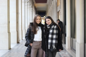 Two women stand side by side in a well-lit, elegant corridor. They are dressed in winter clothing, including coats and a plaid scarf. The corridor features high ceilings, patterned floor tiles, and decorative archways. The setting appears to be indoors, with a warm and inviting atmosphere.