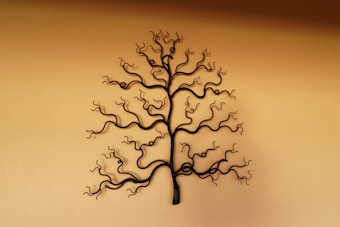 An artistic metal sculpture is mounted on a plain wall, resembling a tree with winding, curly branches. The intricate design of the sculpture presents an abstract and organic form.