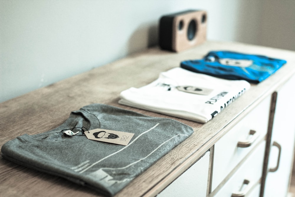 three assorted-color crew-neck shirts folded on top of a wooden dresser