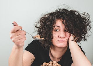 woman with messy hair wearing black crew-neck t-shirt holding spoon with cereals on top