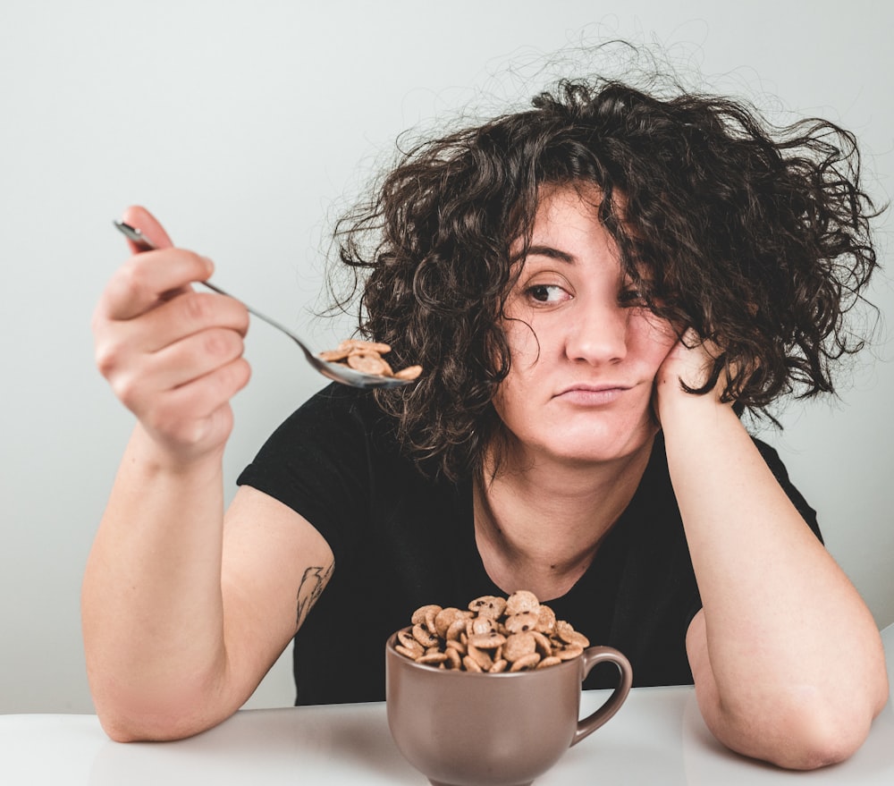 woman with messy hair wearing black crew-neck t-shirt holding spoon with cereals on top