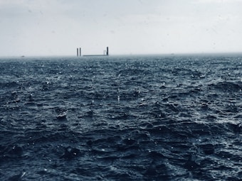 A vast, churning body of water with rough waves and droplets of rain falling onto its surface. In the distance, a small structure with two vertical beams stands amidst a foggy horizon, creating a sense of isolation.