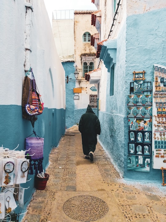person wearing hooded jacket walking on alley between buildings during daytime in Chefchaouen Morocco