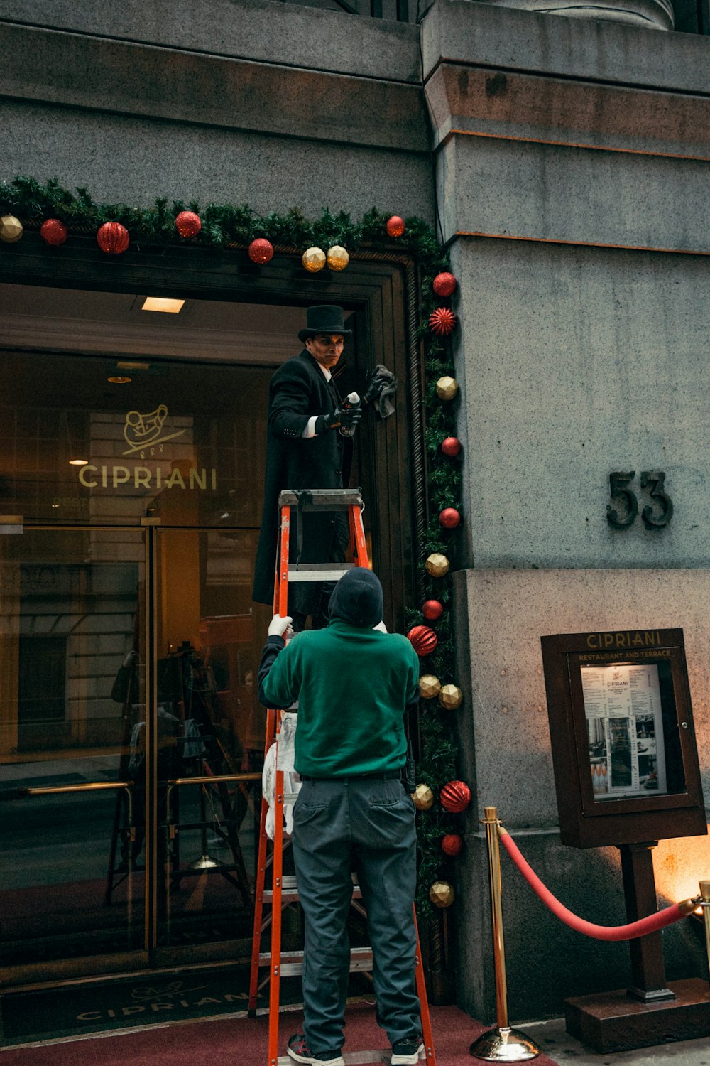 man on top of A-framed ladder being held by another man by glass wall