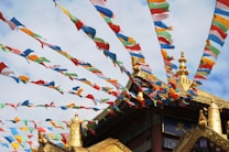 Colorful prayer flags flutter against a backdrop of a traditional temple with intricate golden decorative elements and a partly cloudy sky.