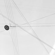 A network of intersecting overhead power lines set against a pale sky, creating a web-like pattern. A single streetlight is visible amidst the crossing wires.