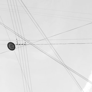 A network of intersecting overhead power lines set against a pale sky, creating a web-like pattern. A single streetlight is visible amidst the crossing wires.