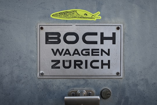 A metallic sign attached to a surface with visible screws displays the words 'BOCH WAAGEN Z&Uuml;RICH'. Above the sign, there is a distinctive yellow, artistic illustration resembling a fish or other aquatic creature.