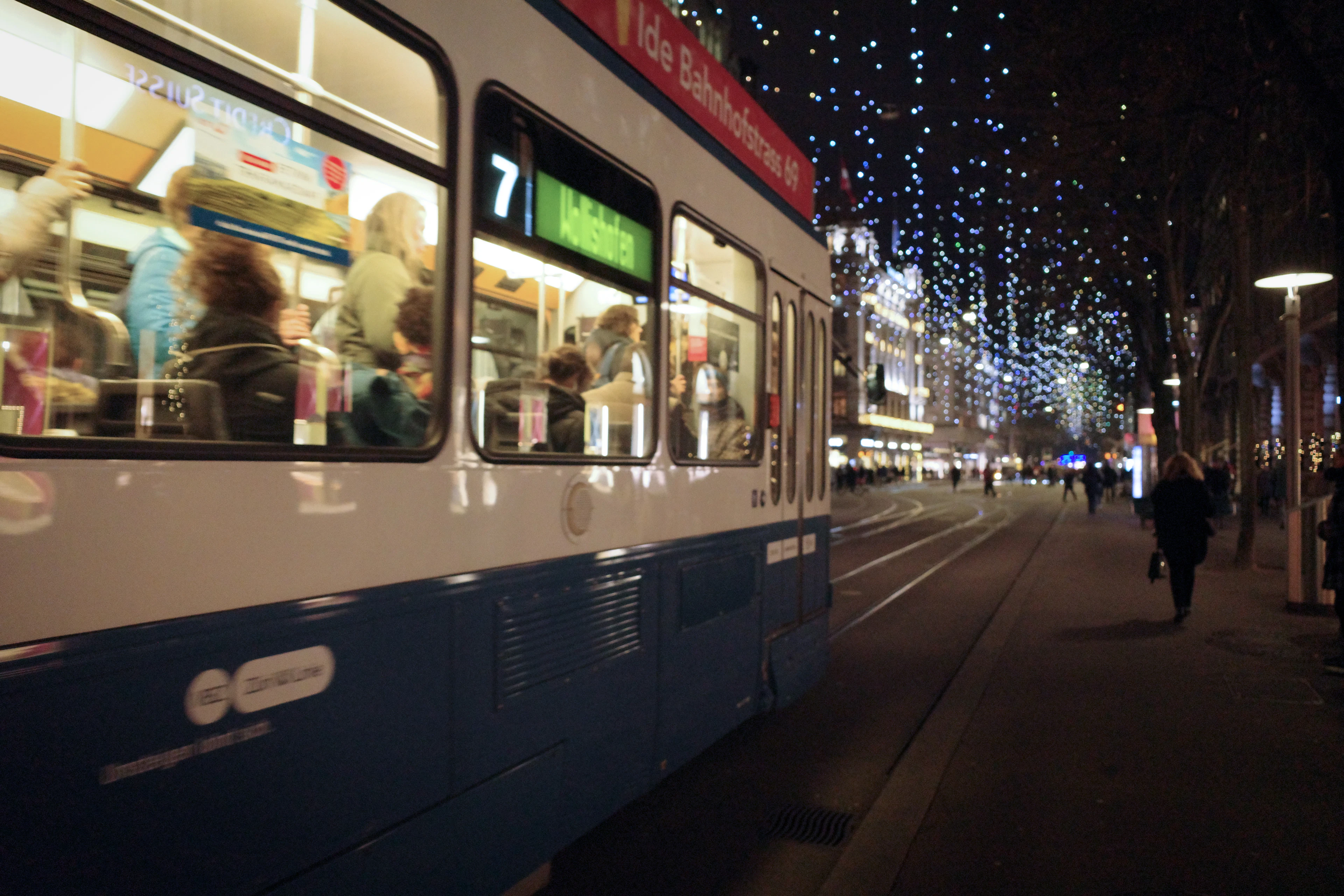 A blue Zurich tram waits on Bahnhofstrasse at night beneath strings of glittering lights.