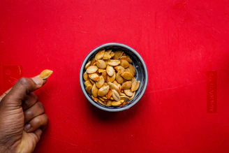 Close-up of golden pumpkin seed oil pouring from a glass bottle with fresh pumpkin seeds scattered around on a dark green background.