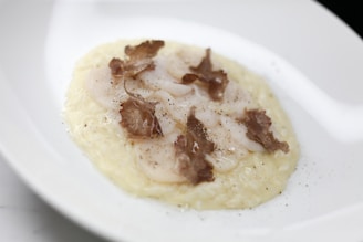A gourmet dish featuring thinly sliced scallops placed on a creamy rice bed, garnished with shaved truffle pieces and a sprinkle of black pepper. The presentation is elegant on a white plate, highlighting the dish's sophistication.