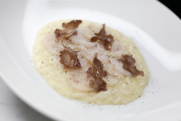 A gourmet dish featuring thinly sliced scallops placed on a creamy rice bed, garnished with shaved truffle pieces and a sprinkle of black pepper. The presentation is elegant on a white plate, highlighting the dish's sophistication.
