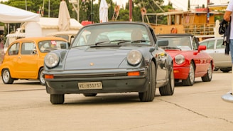 A collection of vintage cars is parked outdoors, featuring a classic black Porsche prominently in the foreground, while a yellow car and a red convertible are visible in the background. The setting appears to be an open area, possibly a car show or exhibition, with tents and people in the vicinity.