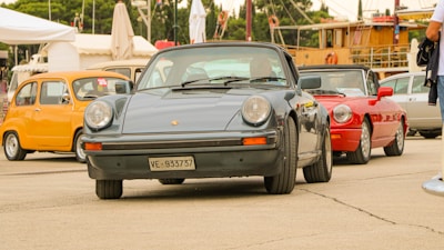 A collection of vintage cars is parked outdoors, featuring a classic black Porsche prominently in the foreground, while a yellow car and a red convertible are visible in the background. The setting appears to be an open area, possibly a car show or exhibition, with tents and people in the vicinity.