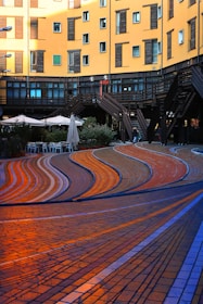 A modern architectural scene features a curved, multi-colored paved walkway with flowing patterns in shades of orange, blue, and brown. Surrounding the path are urban buildings with yellow facades and dark window shutters. There are outdoor seating areas with umbrellas, and a series of wooden staircases offering access to different levels.
