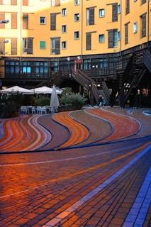 A modern architectural scene features a curved, multi-colored paved walkway with flowing patterns in shades of orange, blue, and brown. Surrounding the path are urban buildings with yellow facades and dark window shutters. There are outdoor seating areas with umbrellas, and a series of wooden staircases offering access to different levels.
