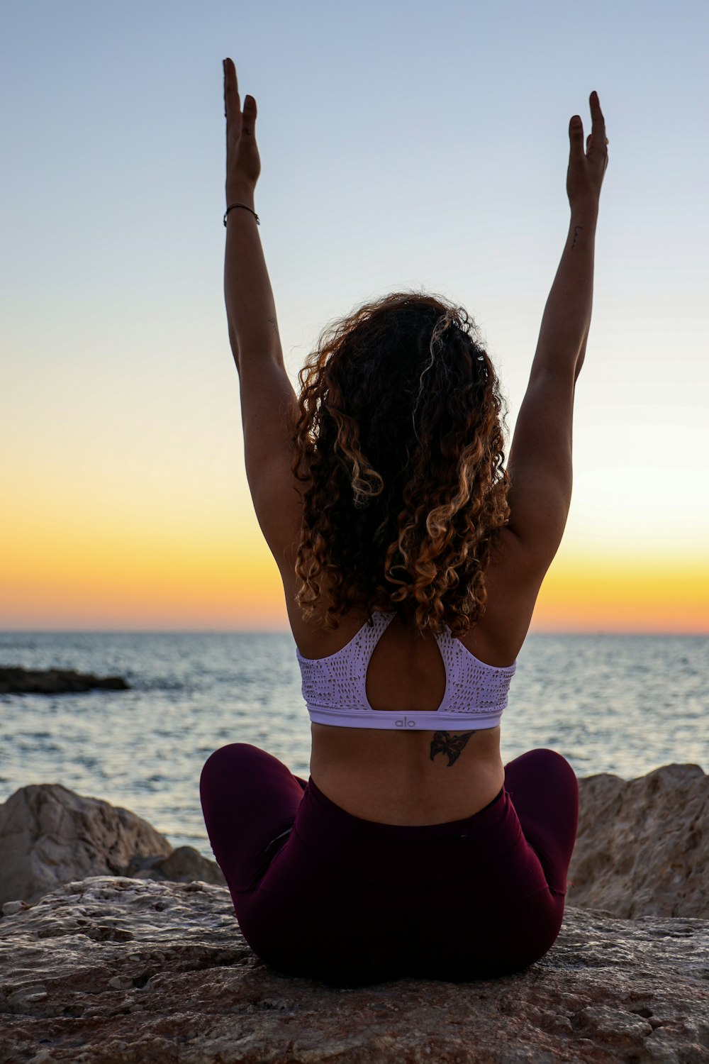 woman raising both her arms while sitting down facing the sea during golden hour