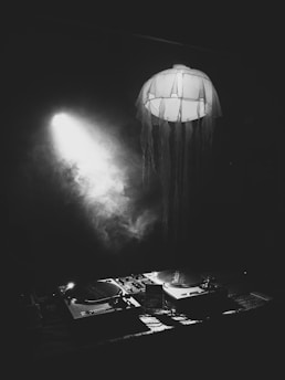 A dimly lit scene with a DJ setup featuring two turntables and a mixer on a table. A spotlight shines from the side, illuminating a jellyfish-shaped hanging decoration. The atmosphere is dark and mysterious, with smoke or fog adding to the ethereal setting.