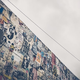 A wall covered with a diverse array of colorful graffiti and posters. Various styles and messages are present, creating a chaotic yet artistic collage. The sky above is overcast, adding a contrast to the vibrant mural.