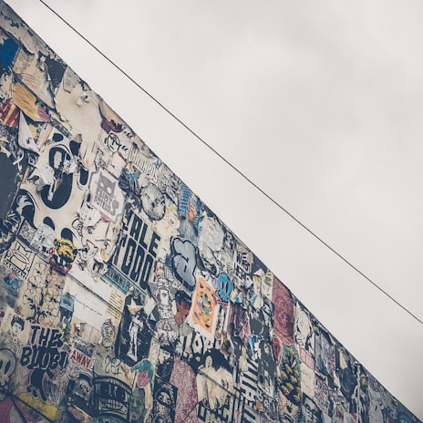 A wall covered with a diverse array of colorful graffiti and posters. Various styles and messages are present, creating a chaotic yet artistic collage. The sky above is overcast, adding a contrast to the vibrant mural.