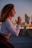 A person is practicing yoga, kneeling with hands in a prayer position. The background features a blurred cityscape with glowing lights, suggesting a sunset or early evening setting.