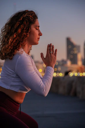 A person is practicing yoga, kneeling with hands in a prayer position. The background features a blurred cityscape with glowing lights, suggesting a sunset or early evening setting.