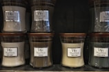 Several glass jar candles with wooden lids are displayed on shelves. The candles have labels with different scents including White Honey, Oudwood, Vanilla Bean, and Evening Bonfire.