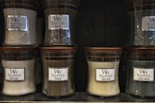 Several glass jar candles with wooden lids are displayed on shelves. The candles have labels with different scents including White Honey, Oudwood, Vanilla Bean, and Evening Bonfire.