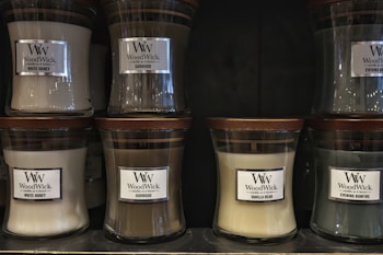 Several glass jar candles with wooden lids are displayed on shelves. The candles have labels with different scents including White Honey, Oudwood, Vanilla Bean, and Evening Bonfire.