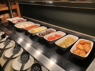 A buffet setup with several trays containing different breakfast items, including sausages, fried eggs, baked beans, scrambled eggs, mushrooms, roasted tomatoes, hash browns, and a potato dish. Serving utensils are laid out alongside the dishes.