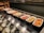 A buffet setup with several trays containing different breakfast items, including sausages, fried eggs, baked beans, scrambled eggs, mushrooms, roasted tomatoes, hash browns, and a potato dish. Serving utensils are laid out alongside the dishes.