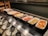 A buffet setup with several trays containing different breakfast items, including sausages, fried eggs, baked beans, scrambled eggs, mushrooms, roasted tomatoes, hash browns, and a potato dish. Serving utensils are laid out alongside the dishes.