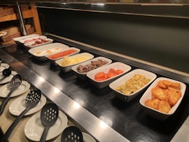 A buffet setup with several trays containing different breakfast items, including sausages, fried eggs, baked beans, scrambled eggs, mushrooms, roasted tomatoes, hash browns, and a potato dish. Serving utensils are laid out alongside the dishes.