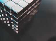 A close-up view of a metallic cube structure, made up of smaller, uniformly sized square blocks with shiny surfaces. The image captures the geometric shapes with a soft blue and gray color tone, and a blurred background.