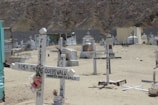 A cemetery scene with simple white wooden crosses marking graves on a sandy terrain. Some crosses have names and dates painted on them, and one has artificial flowers attached. In the background, there are a few larger, more elaborate tombs and crypts. The area appears arid, with a rocky hillside in the distance.