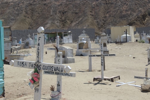 A cemetery scene with simple white wooden crosses marking graves on a sandy terrain. Some crosses have names and dates painted on them, and one has artificial flowers attached. In the background, there are a few larger, more elaborate tombs and crypts. The area appears arid, with a rocky hillside in the distance.