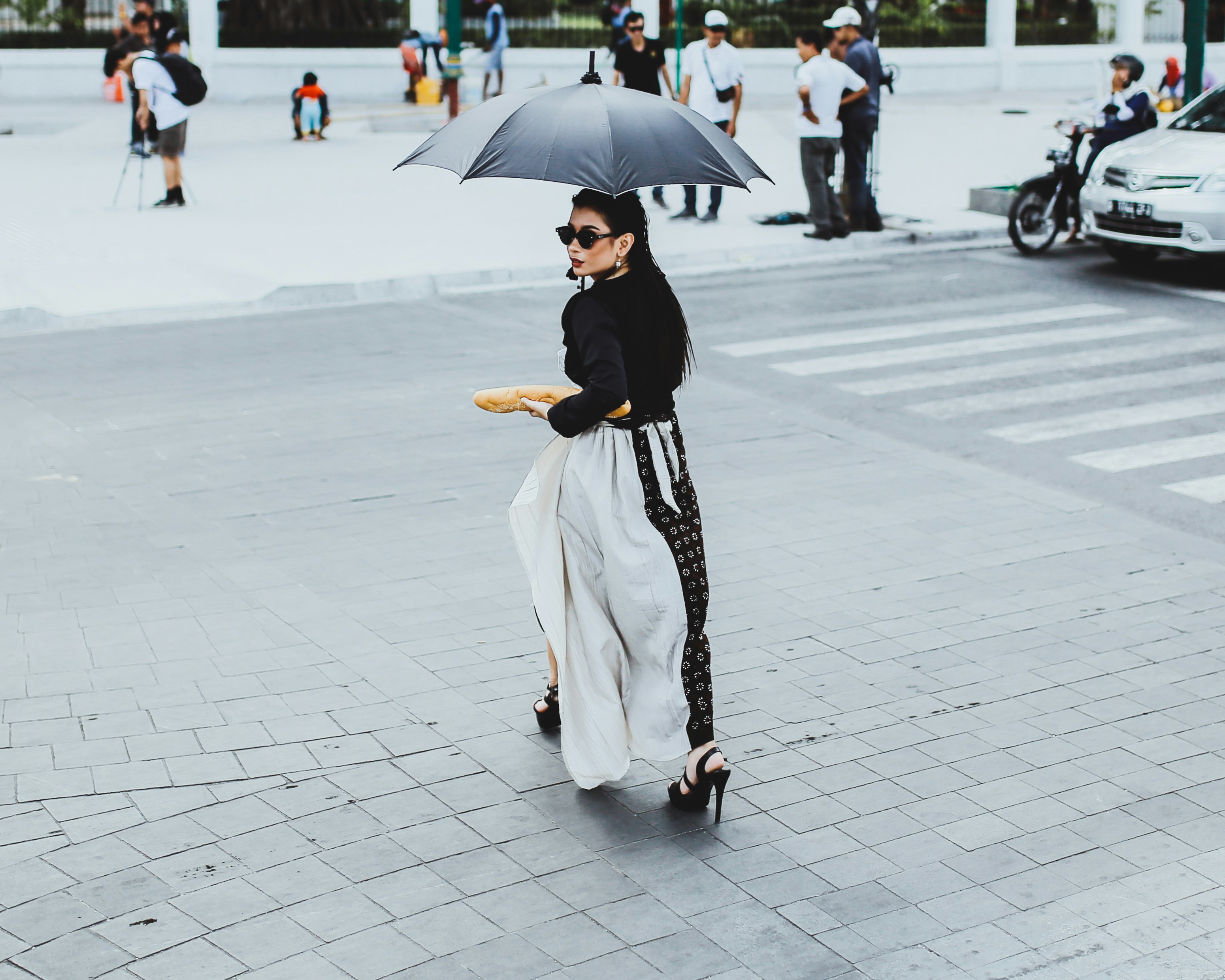 woman wearing black long-sleeved shirt and sunglasses using umbrella while walking on pathway and other people standing 
