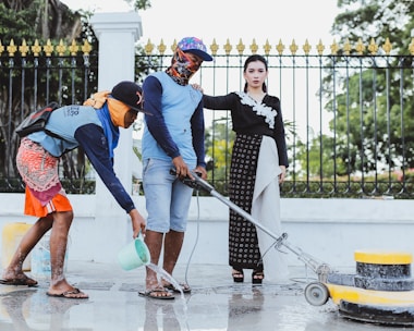 woman standing while holding on shoulder of man cleaning pathway and another man spreading water using green water dipper