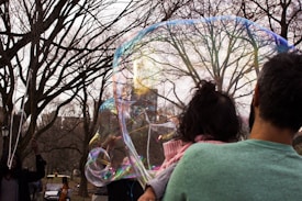A large soap bubble is floating in front of a group of leafless trees, reflecting multiple colors. A person is seen holding a child with their back to the camera, while another individual is raising hands, possibly manipulating the bubble with a stick. The mood is playful and whimsical.