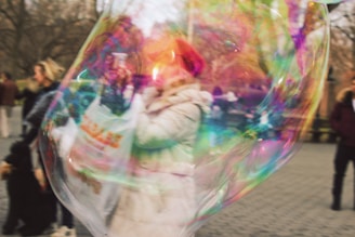 A large, colorful soap bubble partially obscures people in a public outdoor setting. The bubble displays iridescent colors, with a person in a winter coat and red hat visible inside the bubble's distorted reflection.