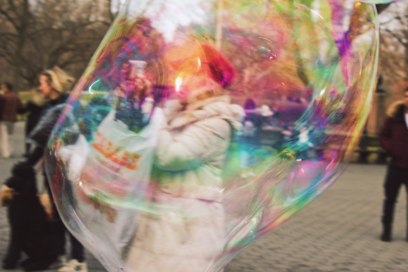 A large, colorful soap bubble partially obscures people in a public outdoor setting. The bubble displays iridescent colors, with a person in a winter coat and red hat visible inside the bubble's distorted reflection.