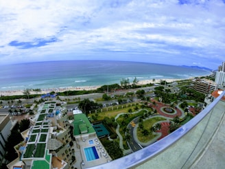 Aerial view of a coastal cityscape featuring a beach with people, a major road with traffic, and numerous residential and recreational buildings. The landscape includes palm trees, a circular park, and swimming pools on top of several buildings. The ocean stretches into the distance under a partly cloudy sky.