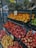 A market stall displaying a variety of fresh fruits, including pineapples, oranges, apples, and peaches. The fruits are arranged neatly in rows on metal racks, with visible price tags. The area is well-lit, and a glass door can be seen in the background, indicating the market's entrance.