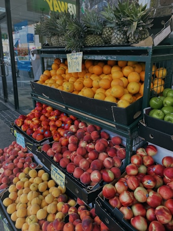 A market stall displaying a variety of fresh fruits, including pineapples, oranges, apples, and peaches. The fruits are arranged neatly in rows on metal racks, with visible price tags. The area is well-lit, and a glass door can be seen in the background, indicating the market's entrance.